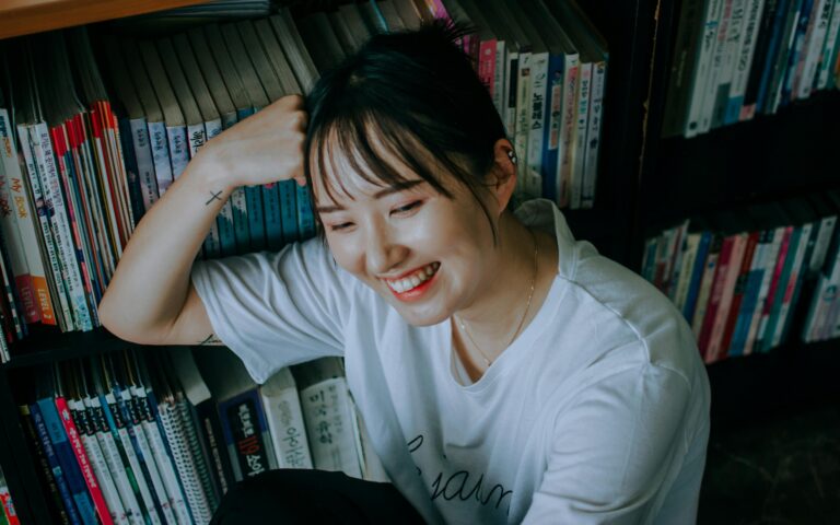 A joyful woman in a white shirt sits and smiles by a bookshelf filled with books.